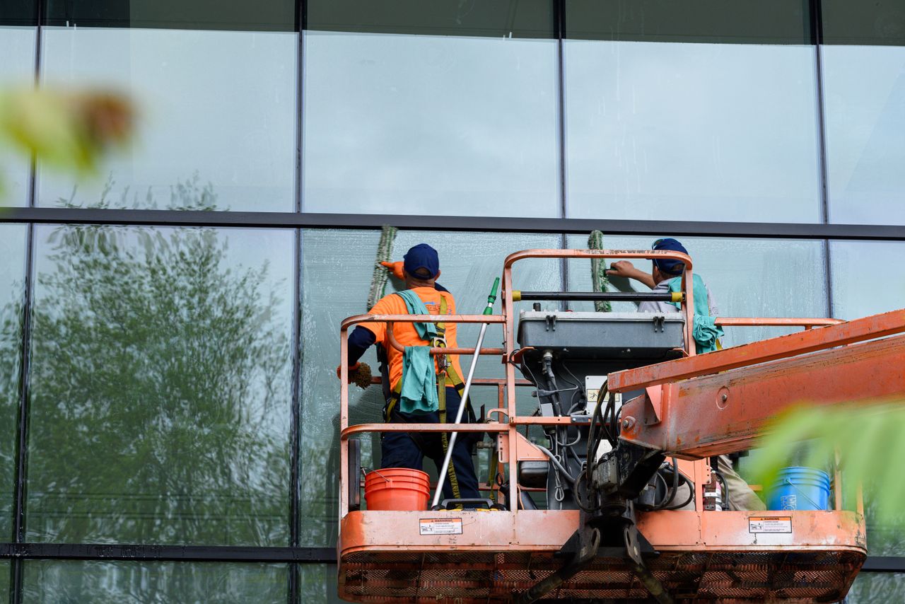 Deux techniciens lavent les vitres d’une façade en verre d’un immeuble à l’aide d’une plateforme élévatrice, afin de valoriser l’esthétique du bâtiment.